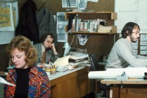 Three people are working in a 1970s-style office filled with papers, books, and architectural drawings. A woman in a patterned jacket sits in the foreground, while two men work at cluttered desks in the background.