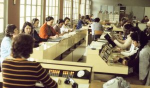 A large room with rows of women working at desks with telecommunication equipment and paperwork. The room is filled with natural light from the windows. Most women are wearing headsets and are focused on their tasks.