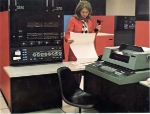 A woman stands in front of an IBM 370 mainframe computer, holding a phone and examining a continuous paper printout. A keyboard and printer are on a nearby desk, with red and black computer cabinets in the background.